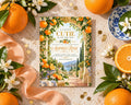 a vibrant invitation card with a garden scene, surrounded by fresh oranges, white flowers, and a blue and white plate.