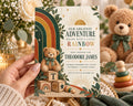 A hand holding a baby shower invitation featuring a teddy bear and a rainbow, with a teddy bear visible in the background.