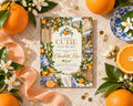 a baby shower invitation with a charming illustration of a market scene, surrounded by oranges, flowers, and a decorative ribbon.