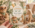 A hand holding a baby shower invitation featuring a cowboy theme with teddy bears, balloons, and baby clothes.