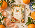 a baby shower invitation with a garden theme, surrounded by oranges, flowers, and a blue and white plate.