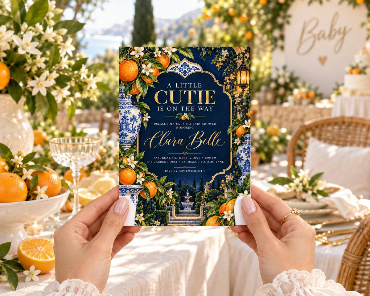 A person is holding a card with a design featuring oranges, flowers, and a garden scene, with the text "A little cutie is on the way" and "Please join us for a baby shower for Clara Belle".