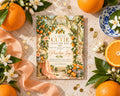 a baby shower invitation with a floral design, surrounded by oranges, white flowers, and a blue and white plate.