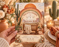 A hand holding a baby shower invitation featuring a desert landscape with cacti, a wooden bench, and a sunburst design.
