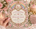 a hand holding a pink and gold invitation card with a rainbow design, surrounded by pink flowers and a pink cake.