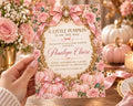 A hand holding a pink and gold invitation card with a pumpkin design, surrounded by pink flowers and pumpkins.