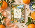 a book titled "A Little Cutie is on the Way" surrounded by oranges, flowers, and a decorative plate.