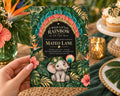 A hand holding a colorful baby shower invitation with a tropical theme, featuring an elephant and lush foliage.