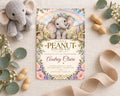 A baby shower invitation featuring a cute elephant surrounded by flowers and greenery, with a ribbon and peanuts on the table.