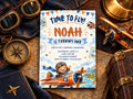 a birthday invitation for a boy named Noah, with a colorful airplane illustration and the text "Time to fly! Noah is turning one". The invitation is surrounded by various objects, including a pair of sunglasses, a compass, and a suitcase, suggesting a travel theme for the boy's birthday party.