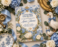 A baby shower invitation featuring a blue and white floral design with illustrations of ducks and flowers, surrounded by decorative elements such as roses, ribbons, and figurines.