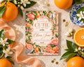 a colorful invitation card with illustrations of oranges, flowers, and a bowl, surrounded by fresh oranges, white flowers, and a blue and white plate.