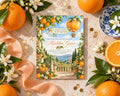 a vibrant invitation card with a garden scene, surrounded by fresh oranges, white flowers, and a blue and white plate.
