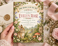 a hand holding a baby shower invitation featuring a rabbit and a garden scene, surrounded by flowers and greenery.